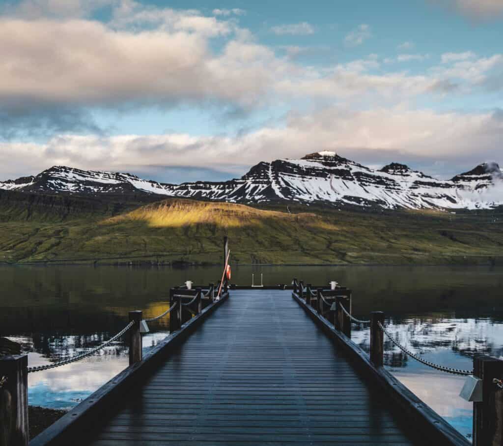 A serene wooden dock leading towards calm waters with snow-capped mountains in the background, a peaceful place to visit in East Iceland.